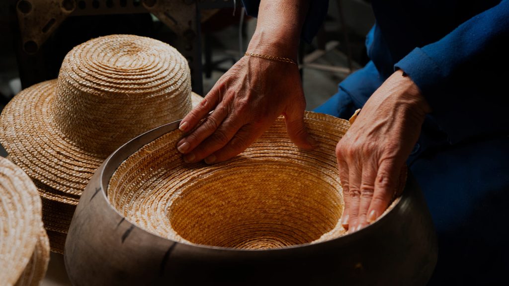 Manos de una artesana dando forma a un cuerpo de sombrero de paja sobre una horma de madera, con más piezas tejidas al lado.