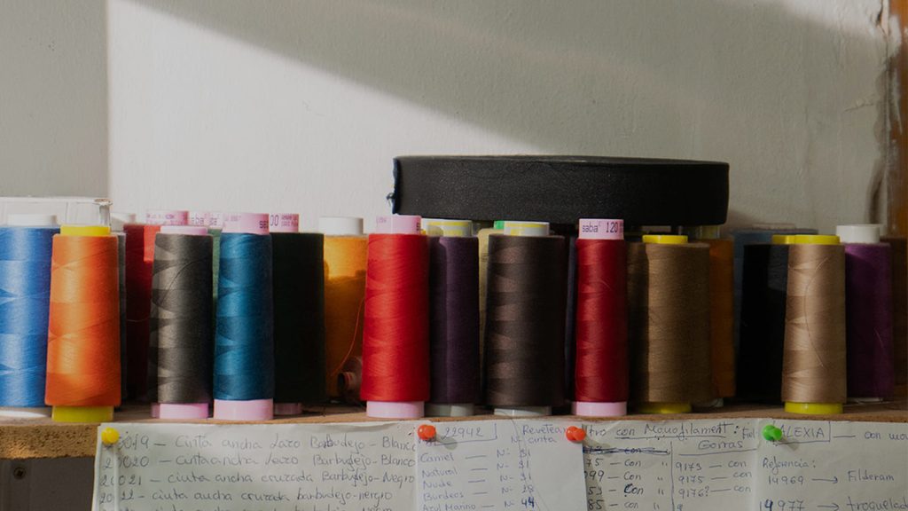 Bobinas de hilo de colores y notas con referencias de cintas en la mesa de costura de un taller de sombrerería artesanal.