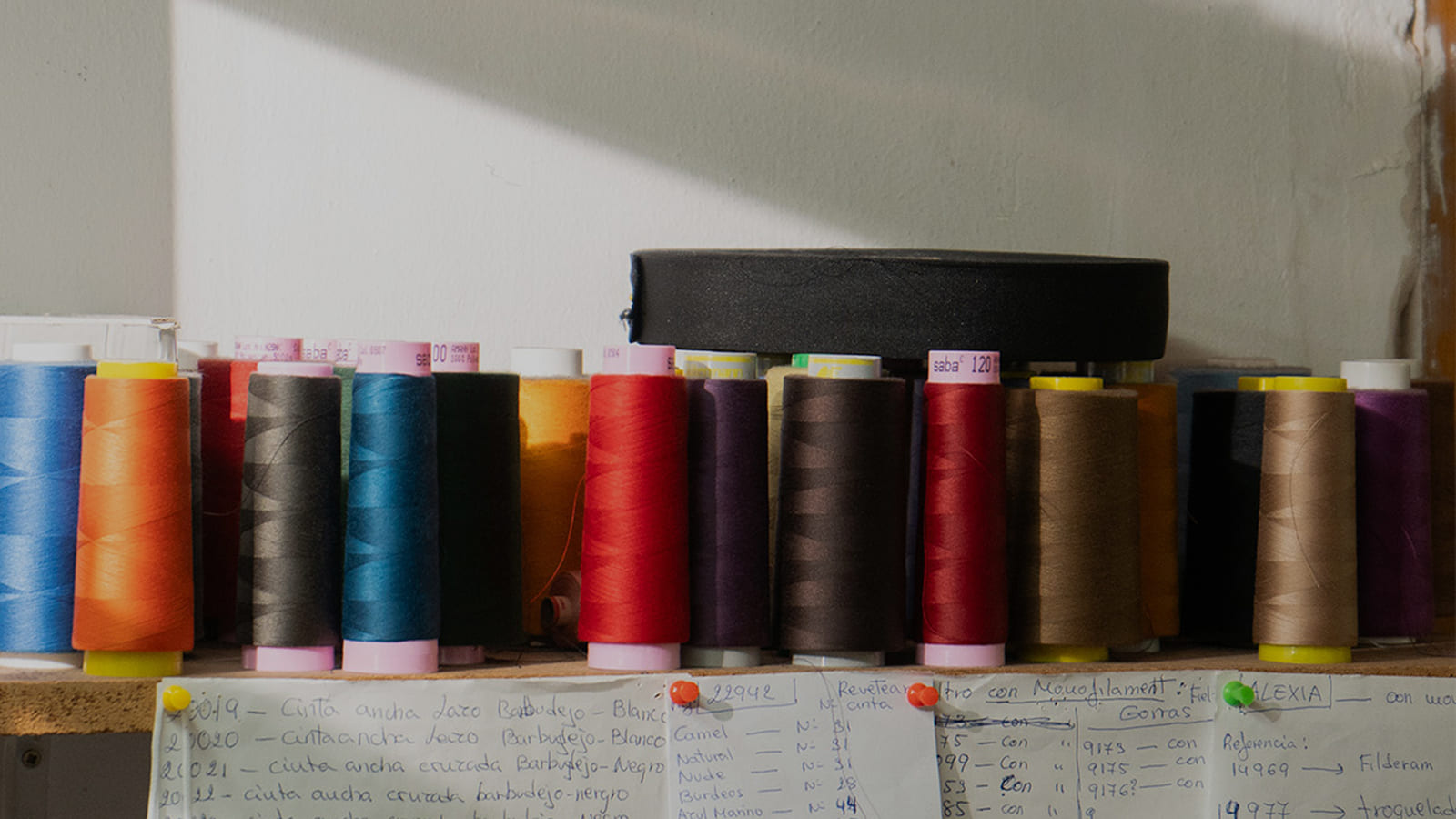Bobinas de hilo de colores y notas con referencias de cintas en la mesa de costura de un taller de sombrerería artesanal.